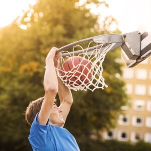 Bilden föreställer en kille som spelar basket. Han håller i basketkorgen samtidigt som basketbollen faller genom nätet. Mål!