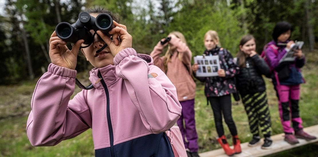 Barn ute i naturen. En flicka som står närmast kameran håller i en kikare.